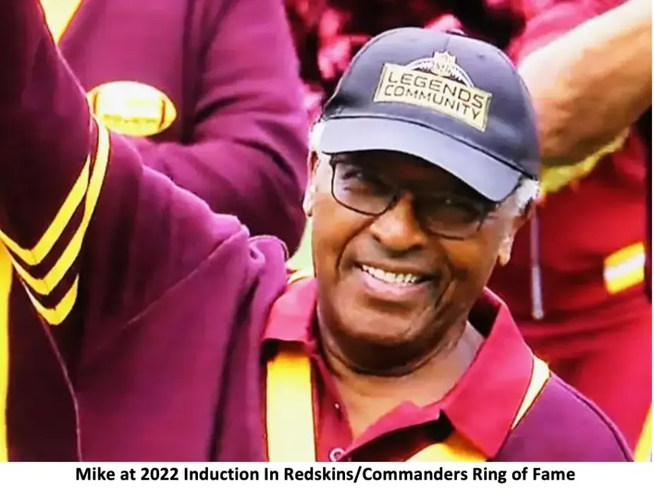 A close portrait of Mike Bass laughing in a dark cap, wearing a burgundy and gold cardigan