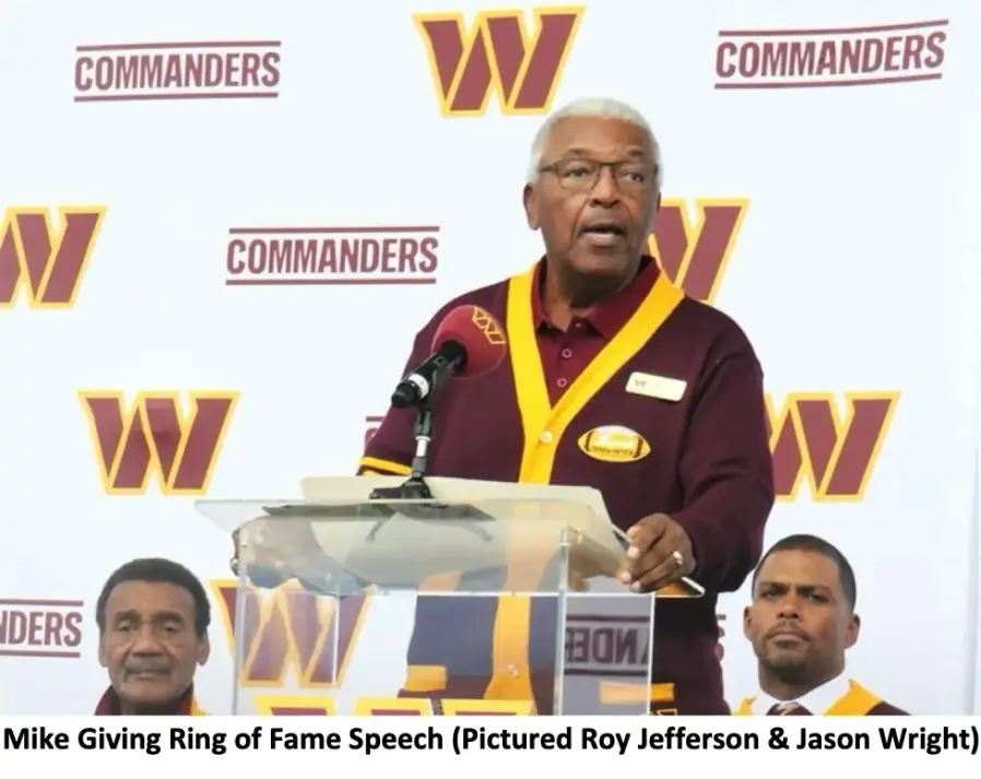 Mike Bass in his Ring of Fame cardigan speaking at a podium with a microphone, in front of a Commanders backdrop