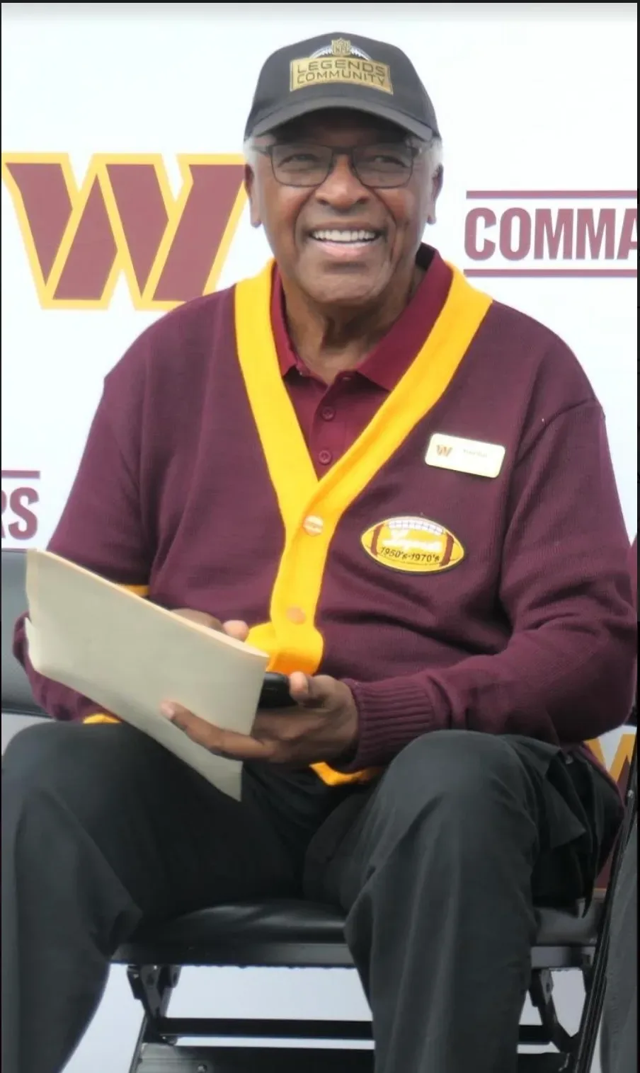 Mike Bass smiling, seated in his Ring of Fame cardigan and Legends Community cap in front of Commanders signage